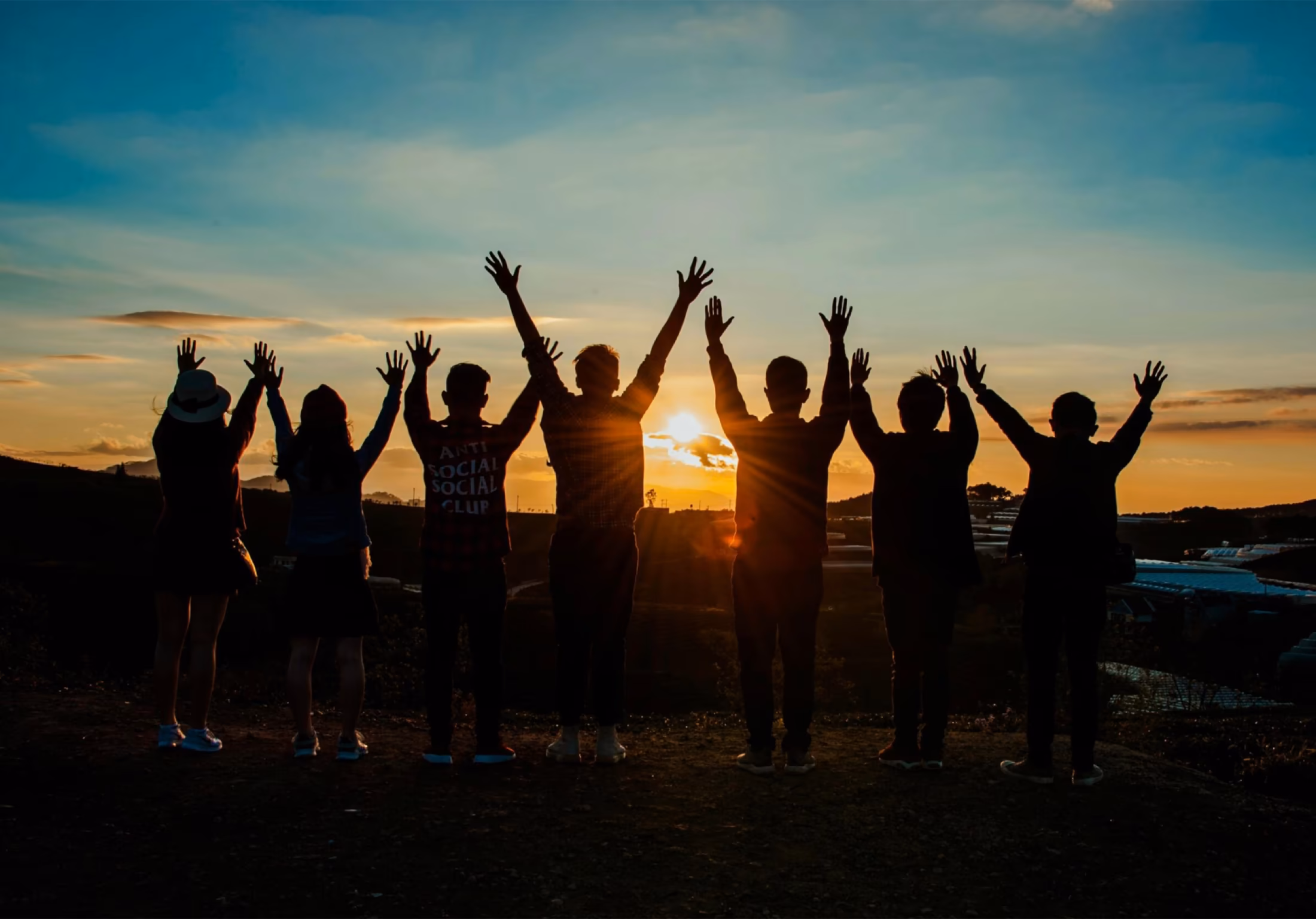 Junge Menschen stehen bei Sonnenuntergang mit erhobenen Händen zusammen.