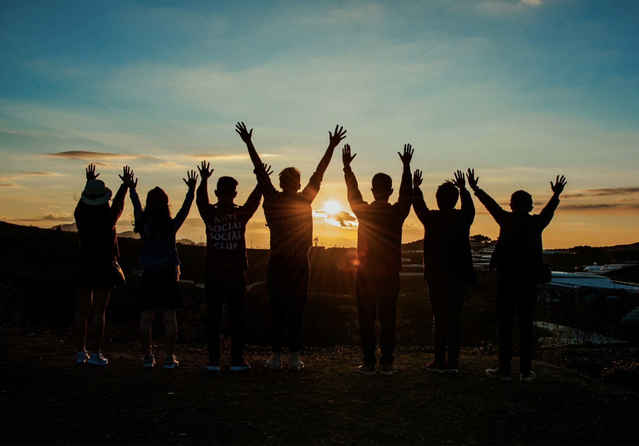 Junge Menschen stehen bei Sonnenuntergang mit erhobenen Händen zusammen.